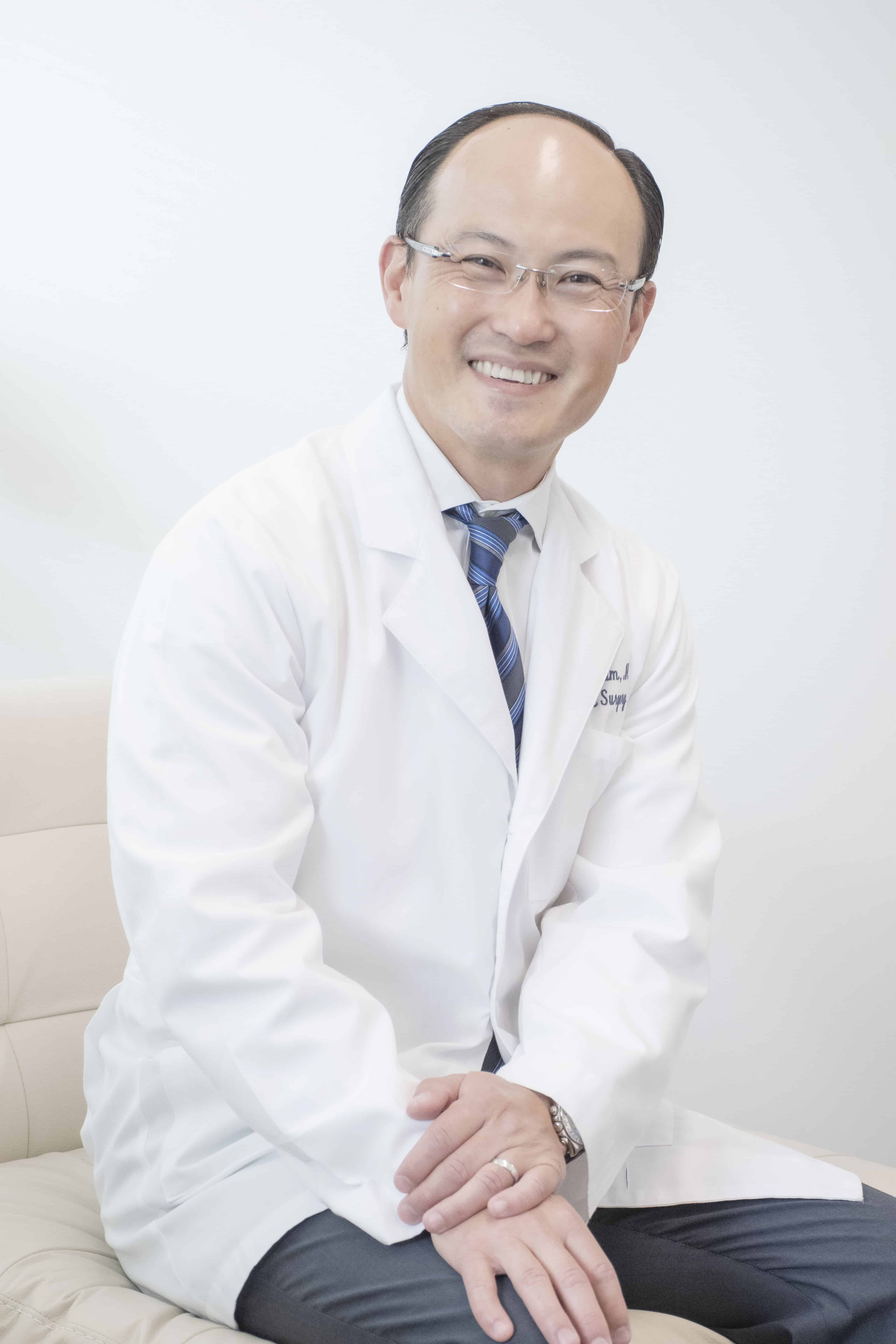A smiling Plastic Surgeon wearing glasses, a white lab coat, a blue striped tie, and a watch sits on a light-colored sofa against a plain, light background in Fairfield County.