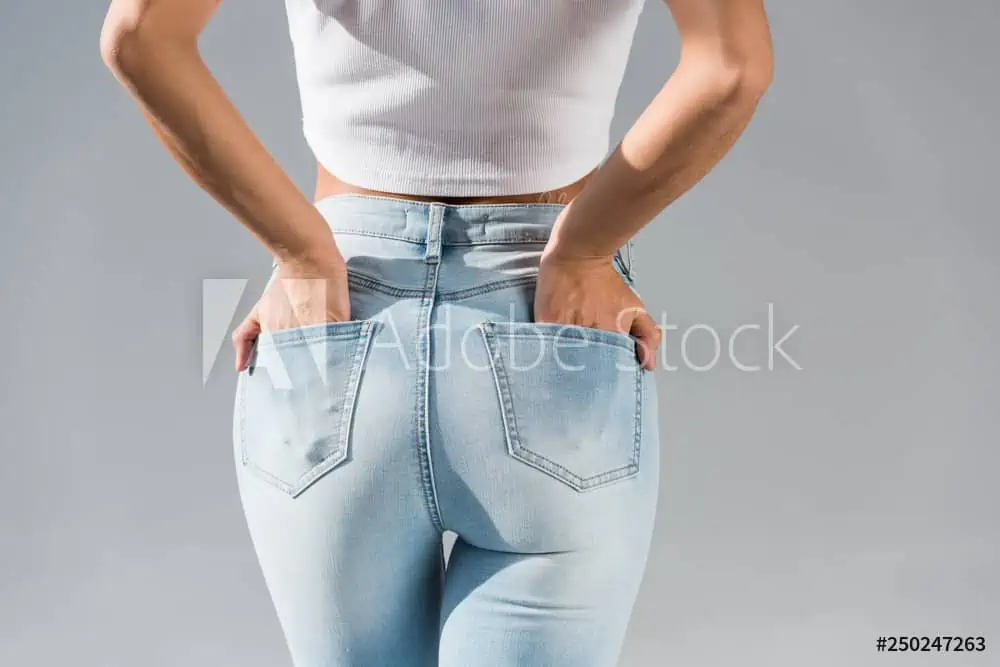 A woman wearing a white crop top and light blue jeans stands with her back to the camera, placing her hands in her back pockets against a plain gray background, showcasing the results of liposuction in Fairfield County.