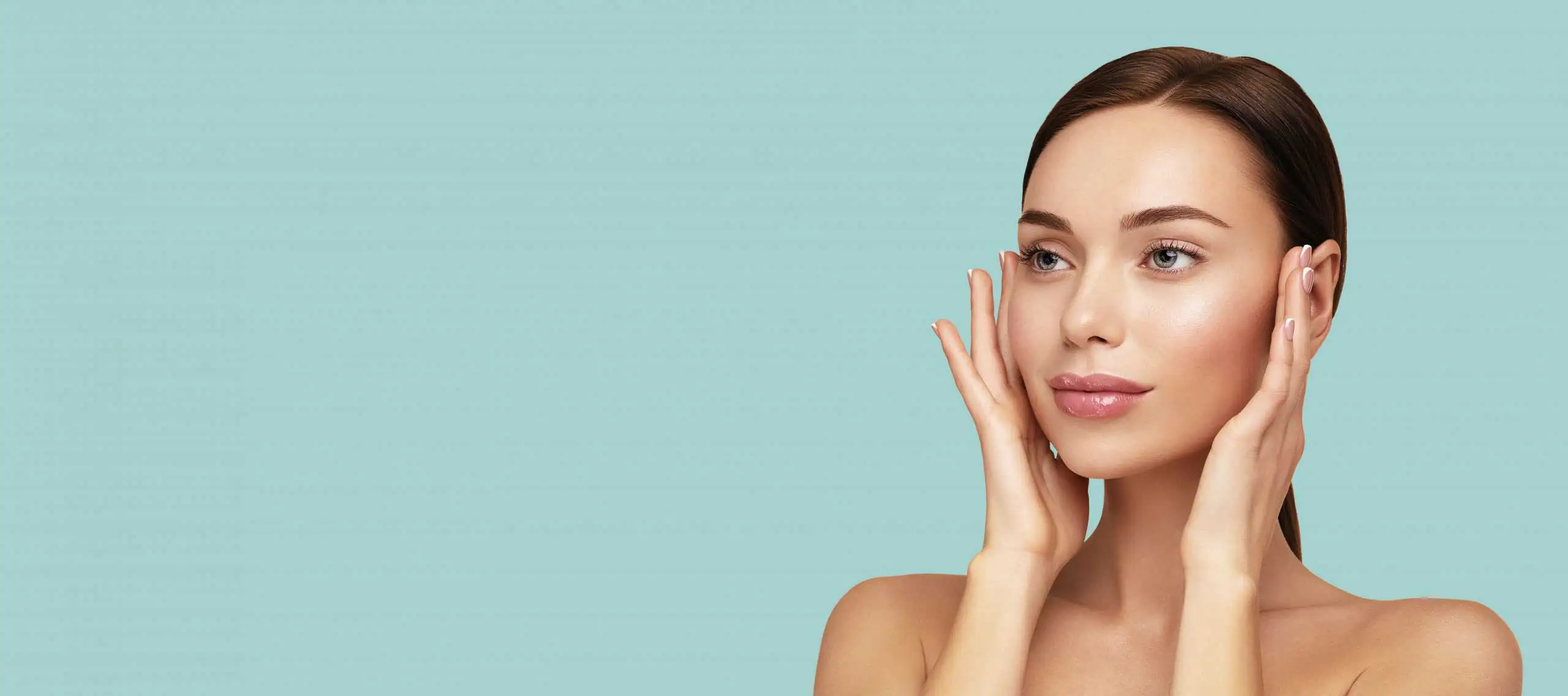 A woman with clear skin touches her face gently with both hands, looking slightly to the side, against a plain light blue background—reflecting the results often achieved with expert care from a Fairfield County plastic surgeon.