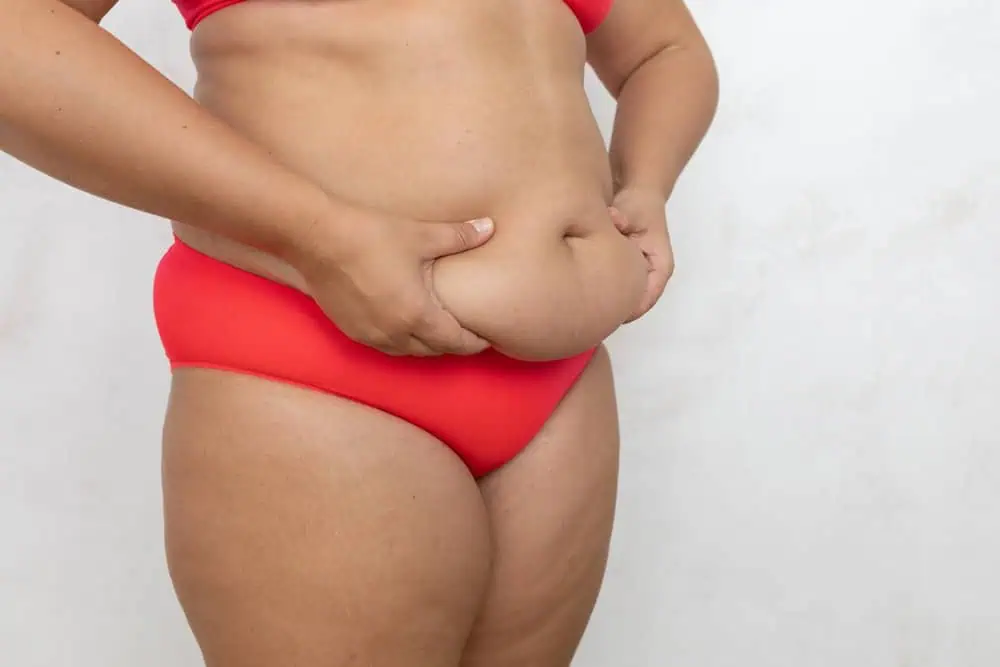 A person in a red bra and underwear stands against a plain background, using both hands to pinch the skin and fat on their stomach.